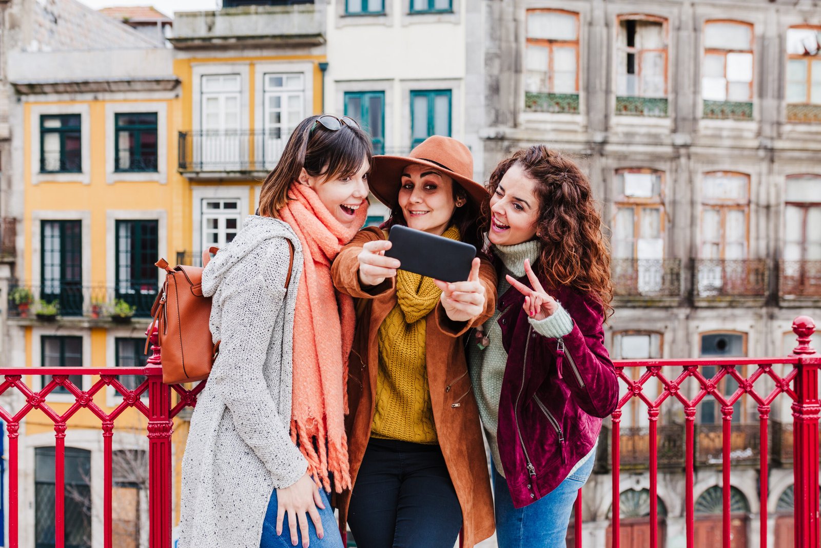 Three female friends taking a selfie on a phone while standing on a bridge against colorful European architecture.